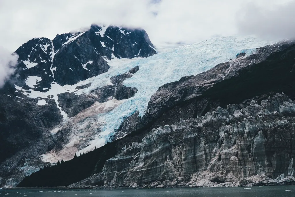 Glacier flowing down a steep mountain into a dark fjord in Kenai Fjords, Alaska