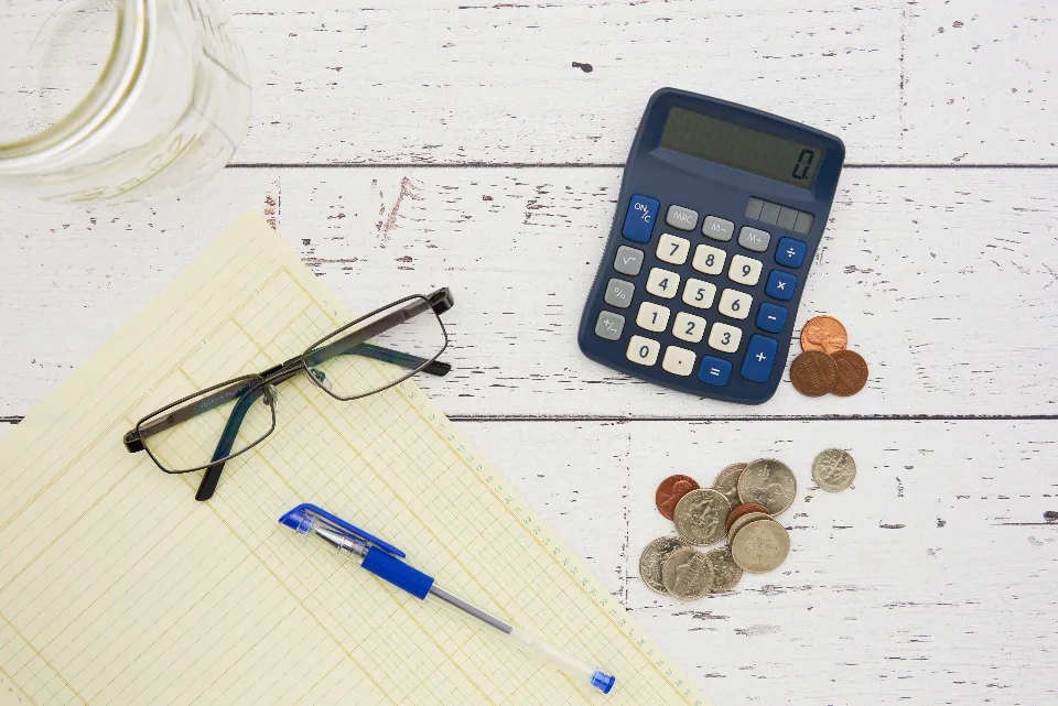 A calculator, U.S. coins, reading glasses and a ledger pad on a white wooden table