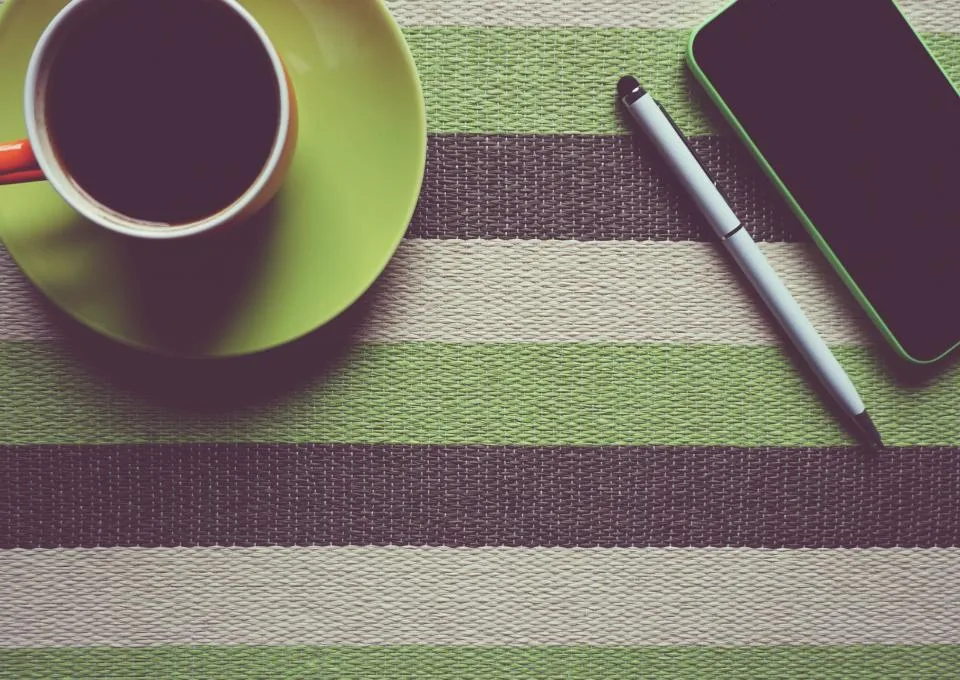 A cup of black coffee on a green saucer beside a pen and a phone on a striped mat