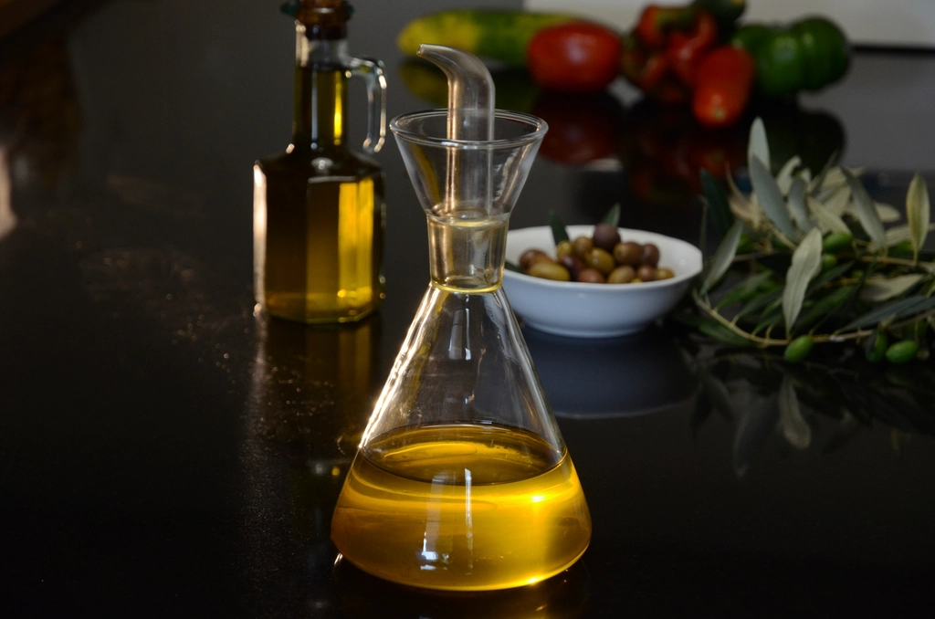 A glass carafe of olive oil beside a bowl of olives and fresh vegetables on a dark counter