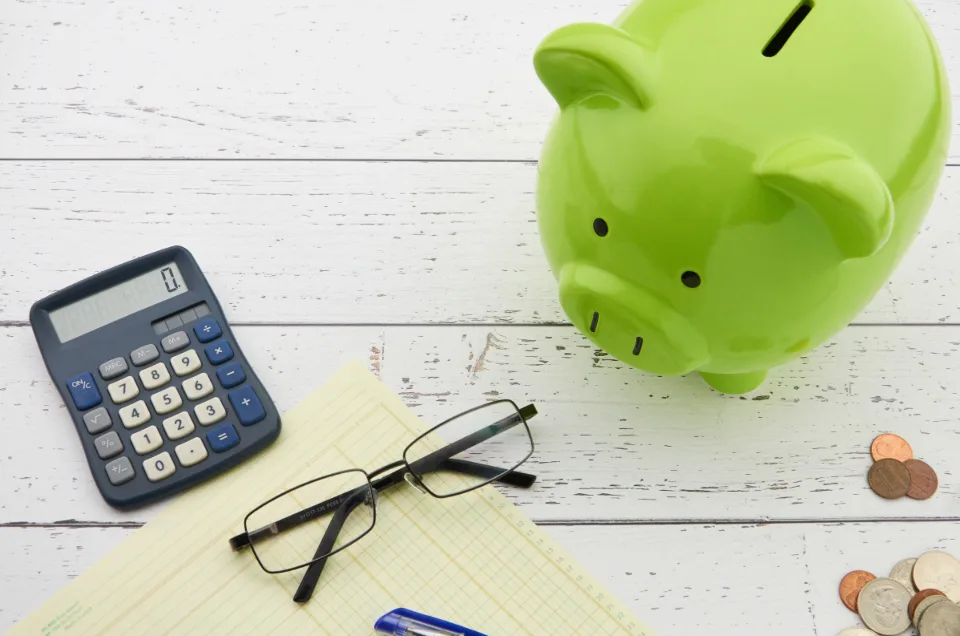 Green piggy bank next to a calculator, glasses and coins on a white wooden table