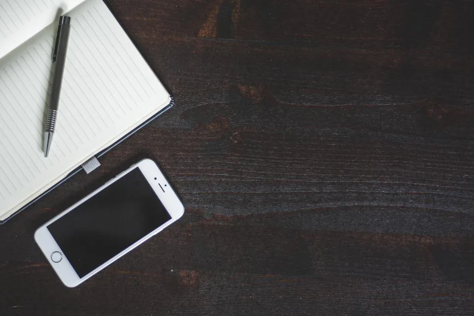 A smartphone with a dark screen lying next to an open notebook and pen on a wooden desk