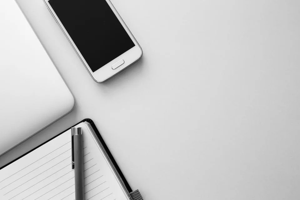 A white smartphone lying on a desk beside a laptop and an open notebook with a pen