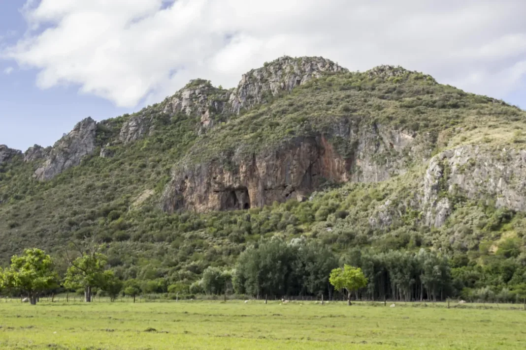 A rocky mountainside with a cave opening above green grassland in South Africa