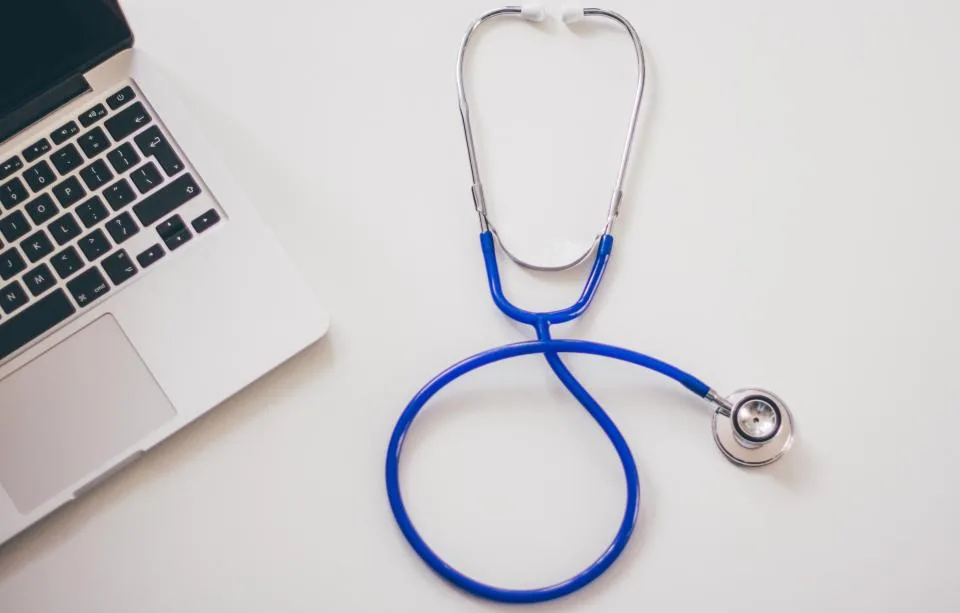 A blue stethoscope on a white desk next to a laptop